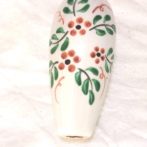May include: A white ceramic bottle stopper with a hand-painted floral design. The design features orange flowers, green leaves, and red swirls. The stopper is approximately 3 inches tall.