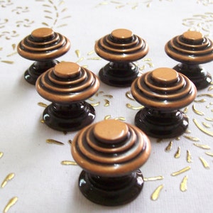 May include: Six copper and black cabinet knobs with a layered design. The knobs have a rounded top and a flat bottom.