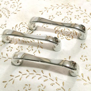 May include: Three silver cabinet door handles with a curved design. The handles are on a white surface with a gold floral pattern.