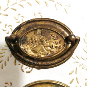 Puede incluir: Un tirador de puerta ovalado de latón con un diseño en relieve de una mujer y un cordero. El tirador tiene un acabado desgastado y antiguo.