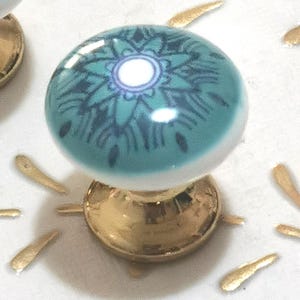 May include: A close-up of a teal and white ceramic cabinet knob with a gold base. The knob features a floral design in dark teal. The base is a shiny gold color. The background is white with gold accents.