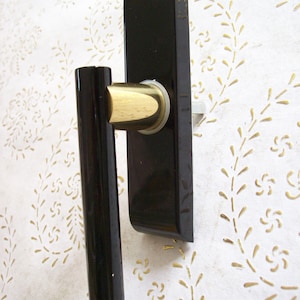 Op de afbeelding: Een zwarte deurklink met een goudkleurig cilindrisch gedeelte en een rechthoekige achterplaat. De klink is gemonteerd op een wit oppervlak met een gouden bloemmotief.