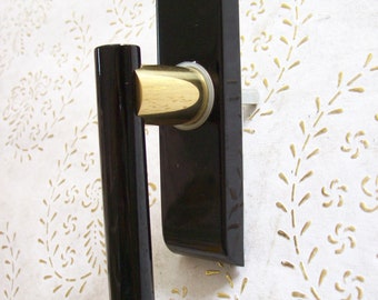 Könnte beinhalten: Ein schwarzer Türgriff mit einem goldfarbenen zylindrischen Abschnitt und einer rechteckigen Rückplatte. Der Griff ist auf einer weißen Oberfläche mit einem goldenen Blumenmuster montiert.