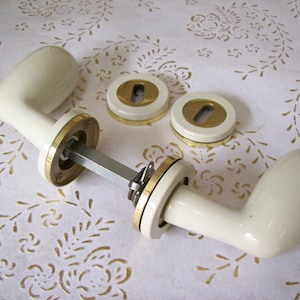 May include: Two white ceramic door handles with gold trim and matching keyhole covers. The handles are oval shaped and have a smooth finish.
