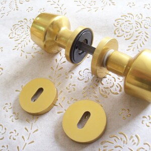 May include: Gold door knob set with two round escutcheons. The knob is cylindrical with a rounded top and bottom. The escutcheons have a keyhole in the centre.