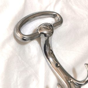 Op de afbeelding: Een glanzende, verchroomde metalen haak met een gebogen handvat en twee onderste haken. De haak heeft een glad, reflecterend oppervlak en wordt waarschijnlijk gebruikt om items op te hangen.
