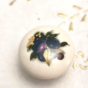 May include: A round, off-white ceramic knob with a detailed illustration of plums, green leaves, and golden accents. The plums are a deep purple with red hues. This decorative knob is suitable for furniture.
