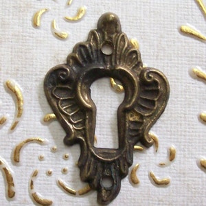 May include: A decorative brass keyhole cover with an ornate design. The cover has a keyhole shape in the center and intricate details around the edges.