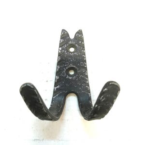 Puede incluir: Un gancho de pared de metal negro con un acabado envejecido. El gancho tiene una forma única con dos brazos curvos y una sección superior con dos agujeros para el montaje. El metal tiene un aspecto texturizado.