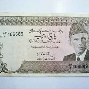 Puede incluir: Un billete de cinco rupias pakistaníes con un retrato de un hombre con sombrero. El billete tiene texto en urdu e inglés, con el número "5" en las esquinas. Número de serie "406689" visible.