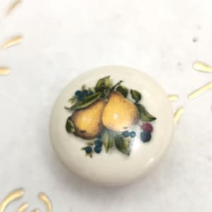 May include: A round, cream-coloured ceramic knob with a detailed illustration of two yellow pears, green leaves, blueberries, and red berries. The knob has a glossy finish, likely for a cabinet or drawer.