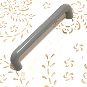 May include: A gray cabinet pull handle with a curved design. The handle is a solid, smooth color and is set against a white surface with a gold floral pattern.