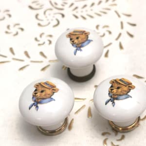 May include: Three white ceramic cabinet knobs with a cartoon bear wearing a hat and blue scarf. The knobs have gold-colored bases and are set against a white background with gold accents. These knobs are ideal for adding a touch of whimsy to furniture.