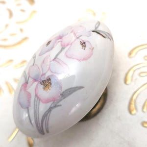 Peut inclure: Bouton de meuble ovale en céramique blanche avec un motif floral rose, doré et gris. Le bouton a une base en laiton et est posé sur une surface blanche avec des accents dorés. Un élément de quincaillerie décoratif.