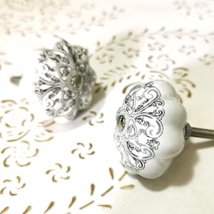 Può includere: Due pomelli per armadi in ceramica bianca con un design floreale bianco e nero. I pomelli hanno una forma arrotondata e rialzata.