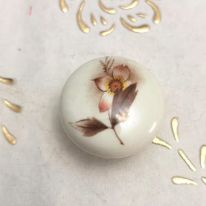 May include: A round, white ceramic knob with a floral design. The flower has orange, pink, and brown hues, with green leaves. The knob is likely for a cabinet or drawer, adding a decorative touch.