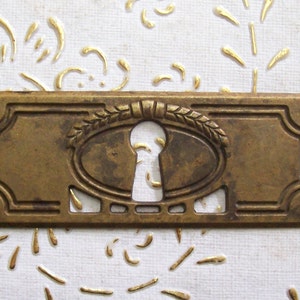 Puede incluir: Una tapa de mirilla de latón con un recorte ovalado y un diseño decorativo de espigas de trigo. La tapa tiene un aspecto ligeramente envejecido, antiguo.