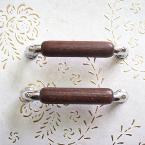 Könnte beinhalten: Zwei braune Holzknaufgriffe mit verchromten Akzenten. Die Griffe befinden sich auf einem weißen Hintergrund mit einem goldenen Blumenmuster.