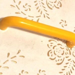 Peut inclure: Une poignée de tiroir incurvée jaune-orangé avec des extrémités arrondies. La quincaillerie est faite d'un matériau lisse et brillant. Le fond est une surface crème avec un motif floral.