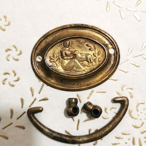 Op de afbeelding: Een set antieke messing kastbeslag met een decoratieve ovale plaat met een vrouw en een lam. De plaat is omgeven door een bloemmotief. De set bevat ook een gebogen handvat en twee knoppen.