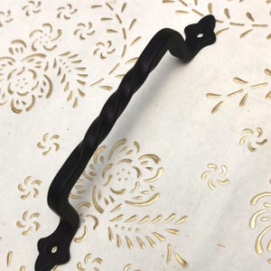 Peut inclure: Une poignée de porte en métal noir torsadé avec des extrémités décoratives. La poignée est conçue pour une armoire ou un tiroir. Le fond est une surface blanche avec un motif floral doré.
