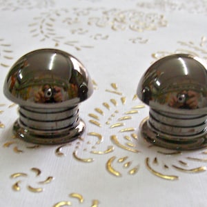 May include: Two black metal bottle stoppers with a shiny, reflective finish. Each stopper has a rounded top and a ridged base.
