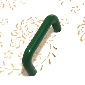 Puede incluir: Un tirador de armario verde curvado con un acabado brillante. El tirador tiene una forma sólida y redondeada, diseñado para un agarre fácil. El fondo es blanco con un estampado floral dorado.