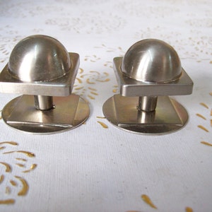 Könnte beinhalten: Zwei silberne Schrankknöpfe mit quadratischer Basis und abgerundeter Oberseite. Die Knöpfe befinden sich auf einer weißen Oberfläche.