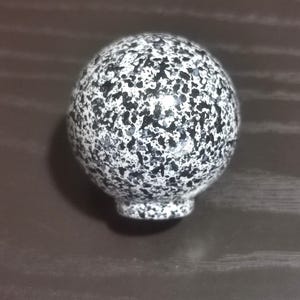 Op de afbeelding: Een bolvormige knop met een gespikkeld patroon van zwarte en witte vlekken over het gehele oppervlak. Het object heeft een gladde, ronde bovenkant en een kleiner, cilindervormig bevestigingspunt aan de onderzijde van de knop.