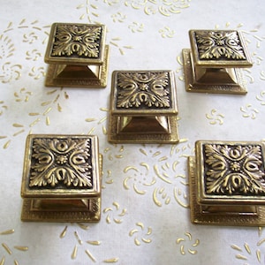 Könnte beinhalten: Fünf goldfarbene Schrankknöpfe mit verzierten Blumendesigns. Jeder Knopf ist quadratisch mit einem erhabenen, detaillierten Blumenmuster.
