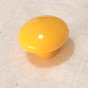 Pode incluir: Um puxador de armário amarelo brilhante, com formato oval. Possui um topo arredondado e uma base cilíndrica curta. A superfície é lisa e brilhante, refletindo a luz. Este hardware é provavelmente para móveis ou armários.