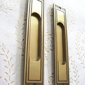 May include: Two gold-toned metal door pulls with a rectangular shape and a recessed handle. The pulls have a decorative border around the edges.