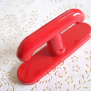 May include: A bright red, vintage-style wooden handle and base. The handle is curved and attached to a rectangular base. The object is likely a tool or a decorative item. The background is a white fabric with a floral pattern.