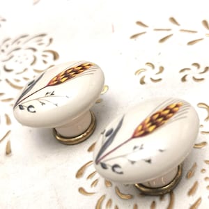 Könnte beinhalten: Zwei ovale cremefarbene Schrankknöpfe. Jeder Knopf hat ein Weizenähren-Design in Gold-, Braun- und Grautönen. Die Knöpfe haben einen goldfarbenen Sockel und sind vor einem weißen Hintergrund mit goldenen Akzenten angeordnet.