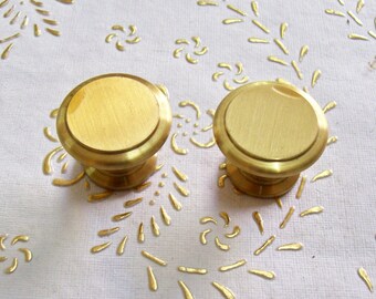 May include: Two gold-toned metal cabinet knobs with a round, slightly raised top and a smooth, polished finish.
