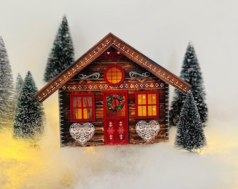 May include: A miniature wooden cabin with a red door and windows, decorated with a wreath and heart-shaped ornaments. The cabin is surrounded by miniature evergreen trees and artificial snow, creating a festive winter scene.