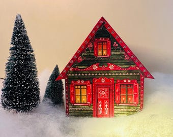 May include: A decorative Christmas house with a red roof and shutters, and a green facade. The house has lit windows and is surrounded by miniature Christmas trees and artificial snow. The house is a festive holiday decoration.