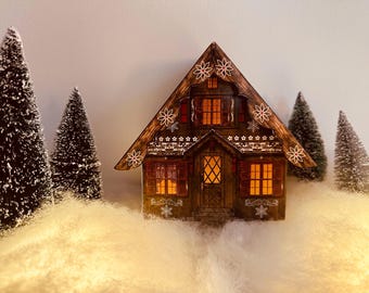 May include: A miniature wooden house with a warm, inviting glow from the windows and door. The house is decorated with snowflake and floral cutouts. The scene includes miniature evergreen trees and a snowy base, creating a cozy winter village scene.