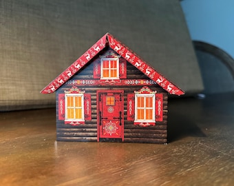 May include: A decorative miniature house with a red roof and accents. The house features a dark brown wood-grain exterior, red shutters, and a red door. The roof is decorated with a white reindeer pattern. The windows have an orange glow.