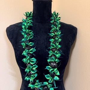 Pu&ograve; includere: Una collana lei verde e nera su un manichino nero. La collana presenta sezioni alternate di perline nere lucide e nastri verdi piegati in modo intricato. Il lei &egrave; una tradizionale ghirlanda hawaiana, con un'estetica tropicale.
