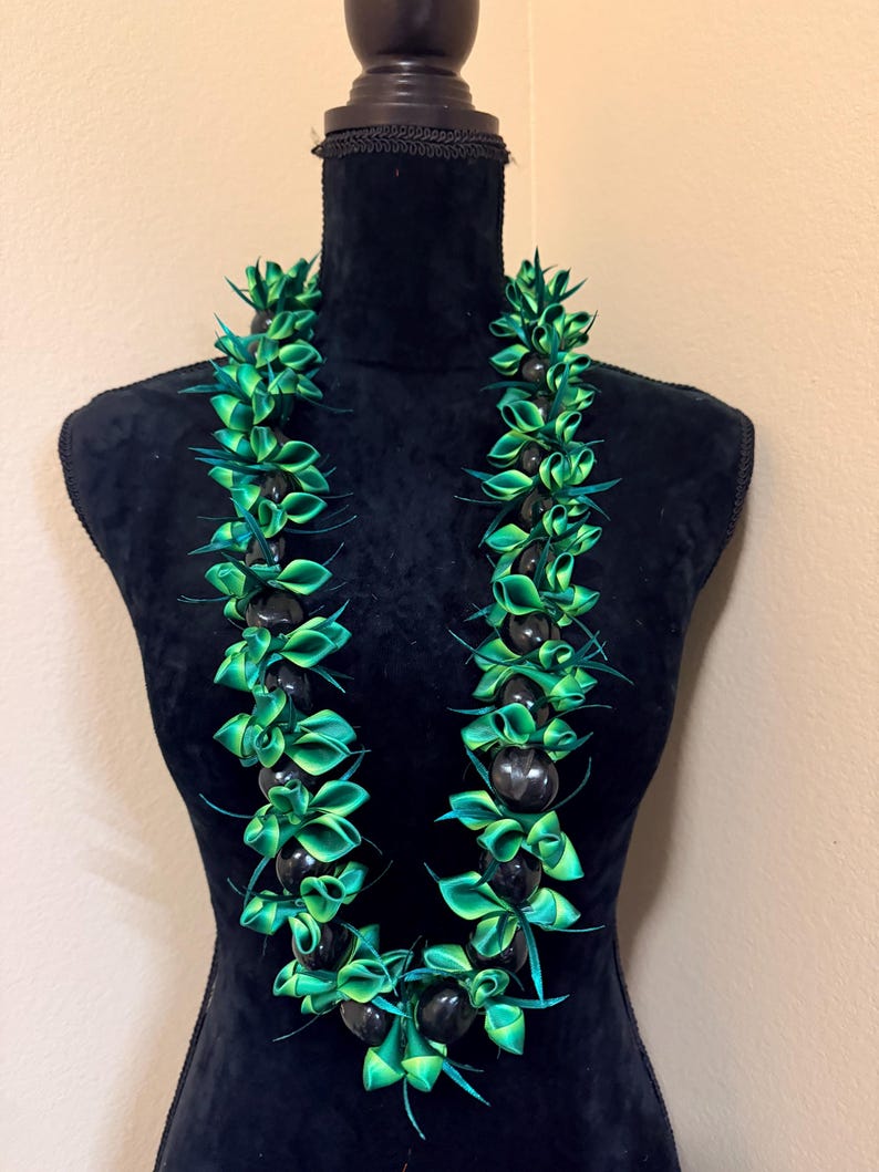 Pu&ograve; includere: Una collana lei verde e nera su un manichino nero. La collana presenta sezioni alternate di perline nere lucide e nastri verdi piegati in modo intricato. Il lei &egrave; una tradizionale ghirlanda hawaiana, con un'estetica tropicale.