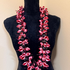 Pu&ograve; includere: Una collana lei con perline scure alternate e petali di fiori rossi e rosa. La lei &egrave; esposta su un manichino nero. I fiori hanno una sfumatura dal rosa al rosso. Una ghirlanda hawaiana tradizionale.
