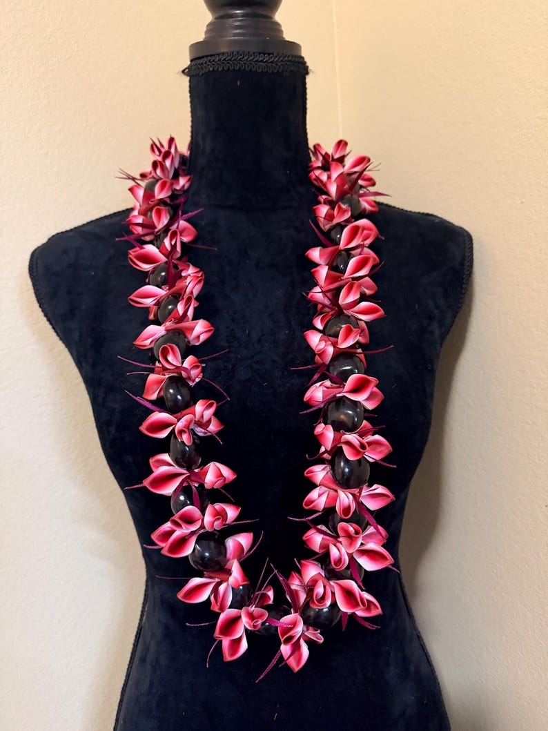 Pu&ograve; includere: Una collana lei con perline scure alternate e petali di fiori rossi e rosa. La lei &egrave; esposta su un manichino nero. I fiori hanno una sfumatura dal rosa al rosso. Una ghirlanda hawaiana tradizionale.
