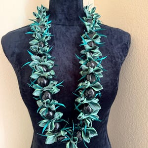 Pu&ograve; includere: Una collana lei esposta su un manichino nero. La lei presenta perline nere alternate e foglie intrecciate verdi e turchese. Le foglie sono disposte in un motivo floreale, creando un'estetica vibrante e tropicale.