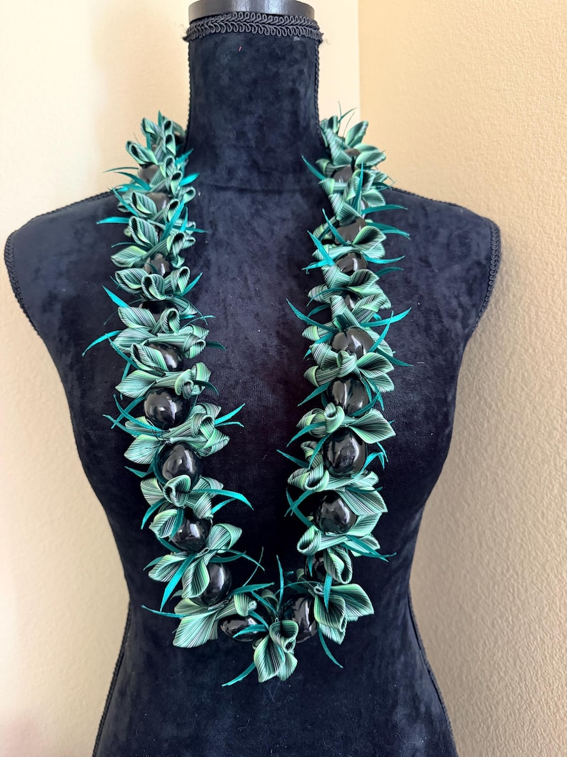 Pu&ograve; includere: Una collana lei esposta su un manichino nero. La lei presenta perline nere alternate e foglie intrecciate verdi e turchese. Le foglie sono disposte in un motivo floreale, creando un'estetica vibrante e tropicale.