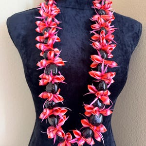 Pu&ograve; includere: Una vivace collana lei esposta su un manichino nero. La lei presenta grappoli alternati di elementi floreali rossi e rosa e perline scure e lucide. Una ghirlanda hawaiana tradizionale.