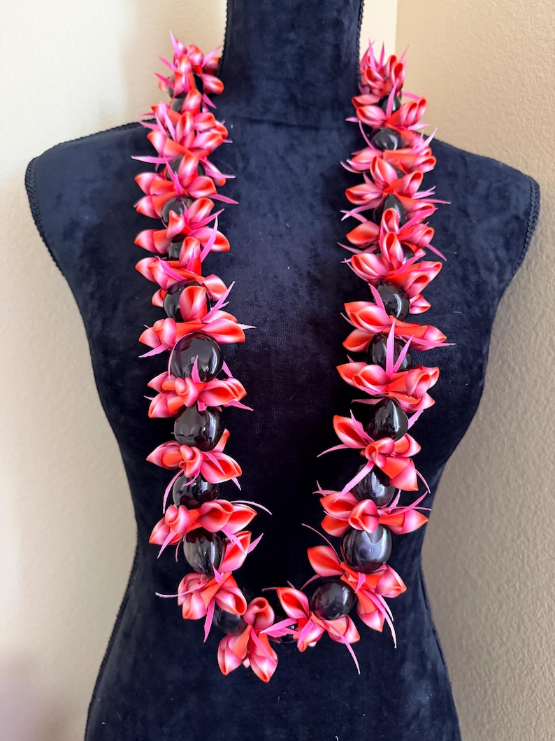 Pu&ograve; includere: Una vivace collana lei esposta su un manichino nero. La lei presenta grappoli alternati di elementi floreali rossi e rosa e perline scure e lucide. Una ghirlanda hawaiana tradizionale.