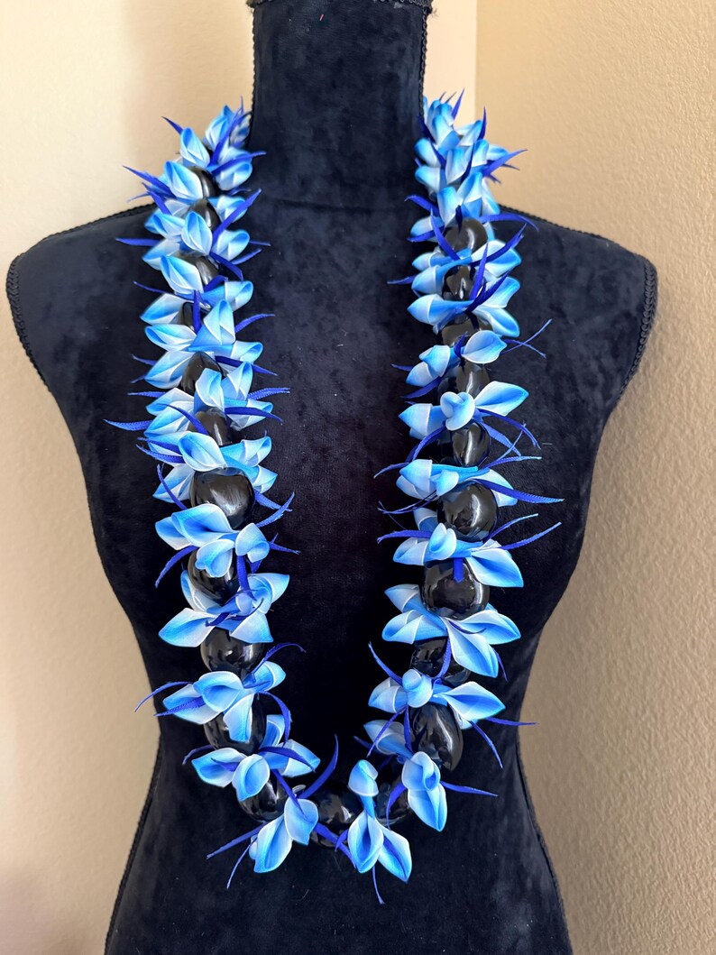 Pu&ograve; includere: Una collana lei con pietre nere alternate e accenti floreali blu e bianchi. La lei &egrave; esposta su un manichino nero, mettendo in risalto i colori vivaci e il design floreale.