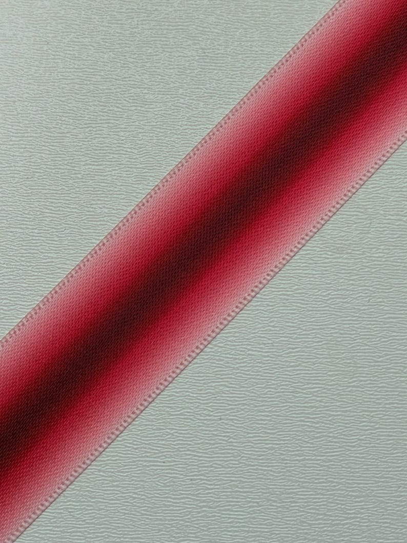 Pu&ograve; includere: Un nastro rosso diagonale con un effetto sfumato, che passa da un bordeaux intenso a un rosa pi&ugrave; chiaro. Il nastro ha un bordo strutturato ed &egrave; posto su uno sfondo grigio chiaro strutturato.