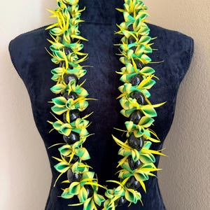 Pu&ograve; includere: Una vivace collana lei con petali a nastro verdi e gialli alternati e perline nere. La lei &egrave; esposta su un manichino nero, mettendo in risalto il suo design colorato e il tema tropicale.